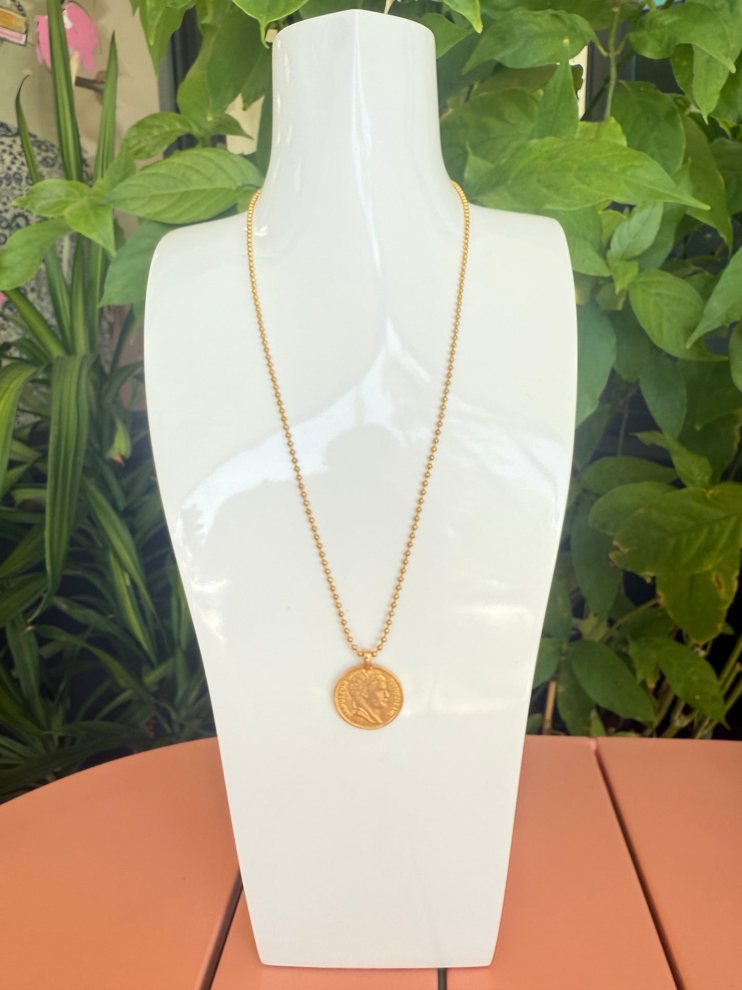 Gold necklace on a white mannequin bust with green foliage in the background. Beaded Necklace with a Coin Charm, 