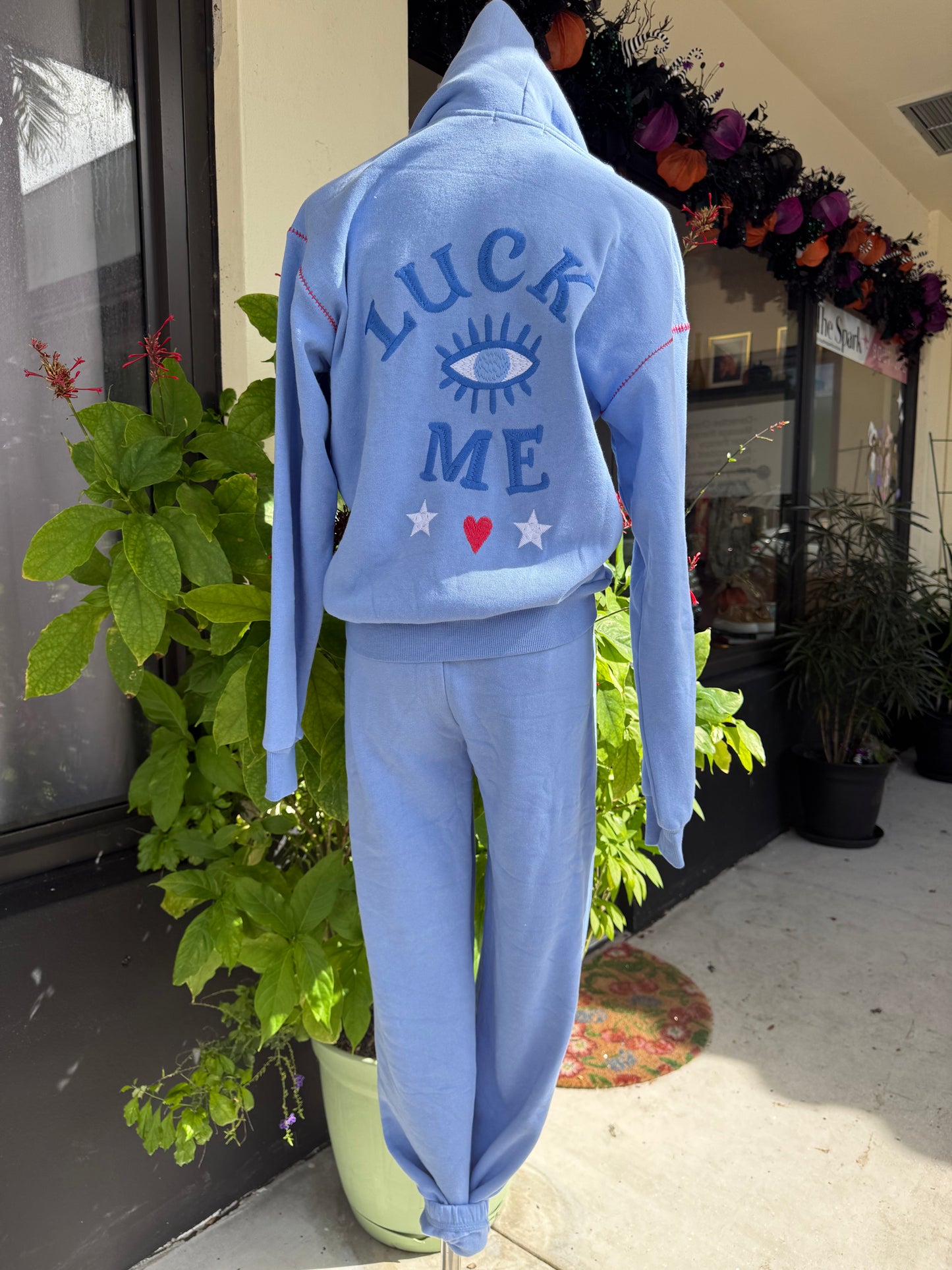 Light blue hoodie with text and graphics on a plant, Blue tracksuit on a mannequin with a decorative background. a staple for curated boutique fashion and chic affordable style available at your Womens Boutique West Palm Beach, FL.
