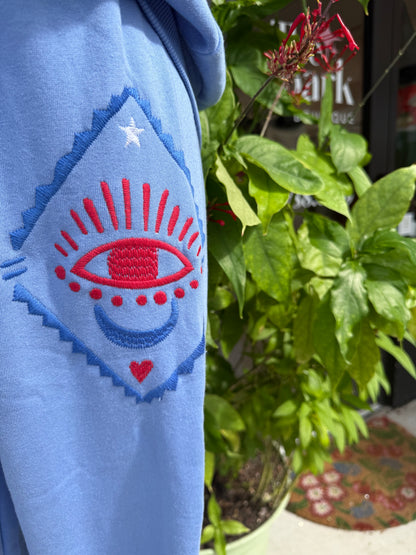 Blue fabric with embroidered design featuring a red heart and blue crescent moon, surrounded by green plants. Blue tracksuit on a mannequin with a decorative background. a staple for curated boutique fashion and chic affordable style available at your Womens Boutique West Palm Beach, FL.

