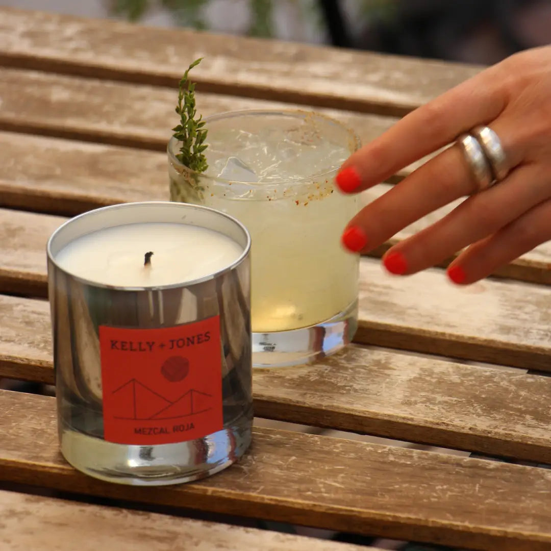 Candle and cocktail on a wooden table with a hand reaching out. a staple for curated boutique fashion and chic affordable style available at your Womens Boutique West Palm Beach, FL.
